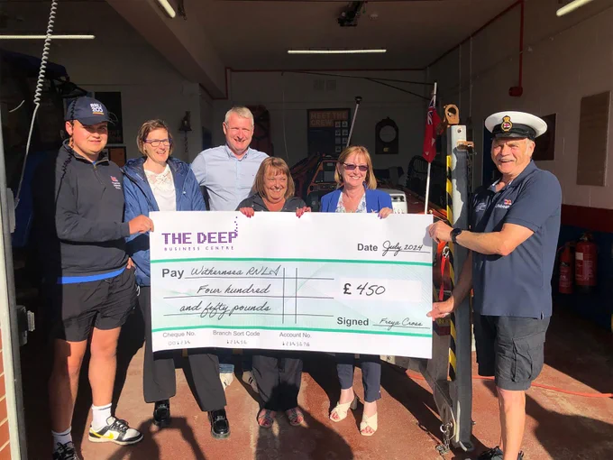 Crew members from the RNLI and staff from The Deep Business Centre all stood smiling and holding a giant cheque for a total of £450.