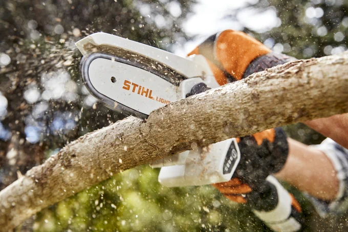 Close-up of a person using a Stihl chainsaw to cut through a thick branch, with wood chips flying from the point of contact.