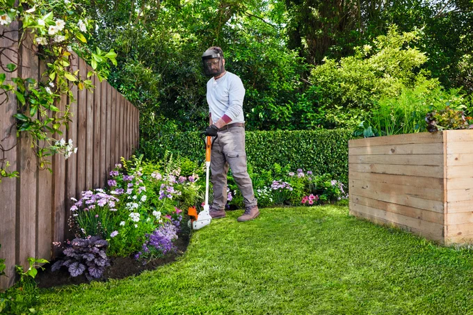 A person in protective gear using a weed trimmer in a lush garden with vibrant flowers and a wooden planter box.