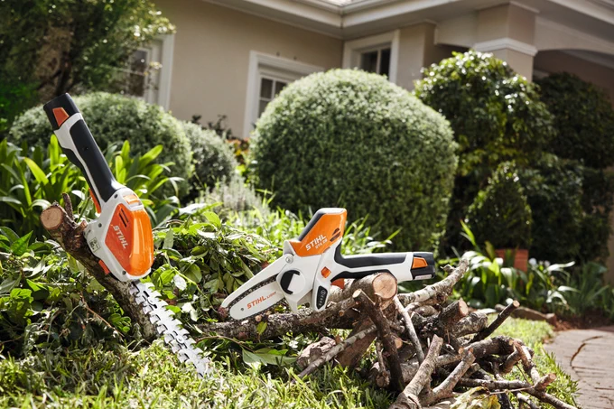 Two chainsaws lying on a pile of cut branches in the front yard of a house with lush greenery.