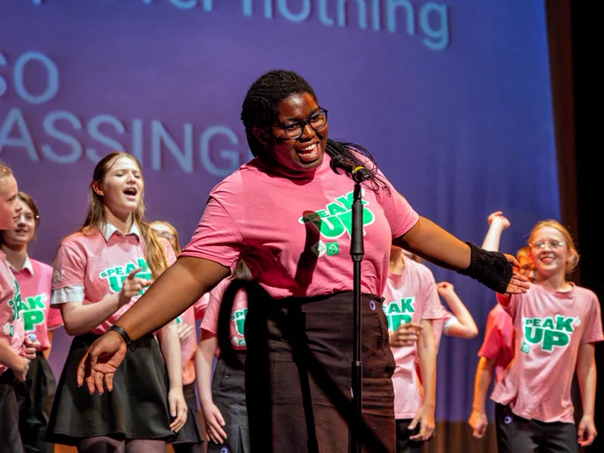 A person in glasses and a pink t-shirt gestures happily during a public speaking event, standing in front of a group of children wearing matching shirts on stage.