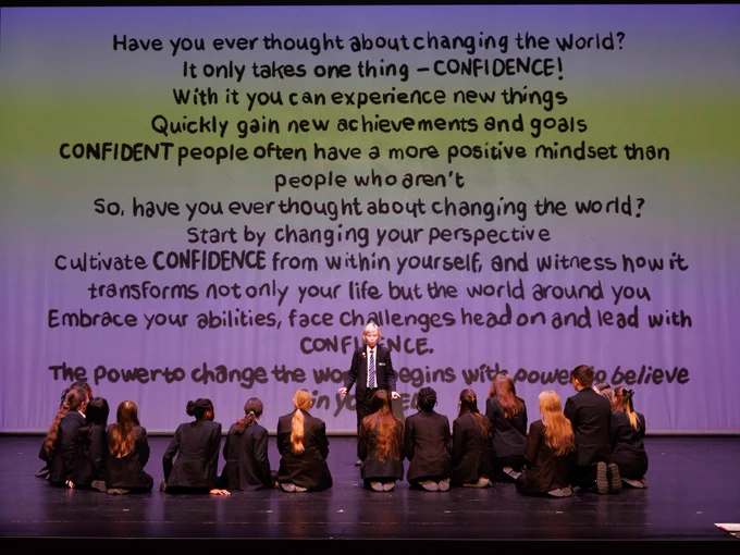 A group of individuals is seated on a stage, viewed from behind, facing a large screen displaying a motivational message about confidence and changing the world. The text emphasizes personal growth and facing challenges with confidence.