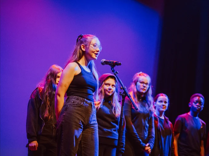 A group of young performers, with one singer taking the lead at the microphone, stands on a stage with ambient blue and purple lighting during a live performance.