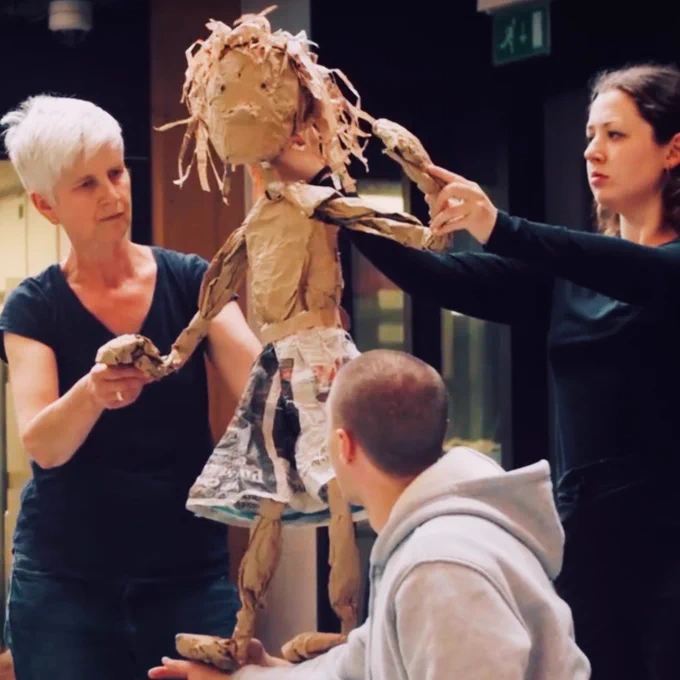 A puppet of a girl made from paper and cardboard looks out from the centre of the image. Two puppeteers each hold one of the puppets arms and head. The puppet is standing on the arms of a dancer who is crouching underneath her. 
