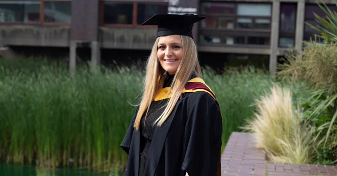 A photo of Dayna smiling at the camera in her graduation cap and gown, standing in front of reeds by the Barbican lake