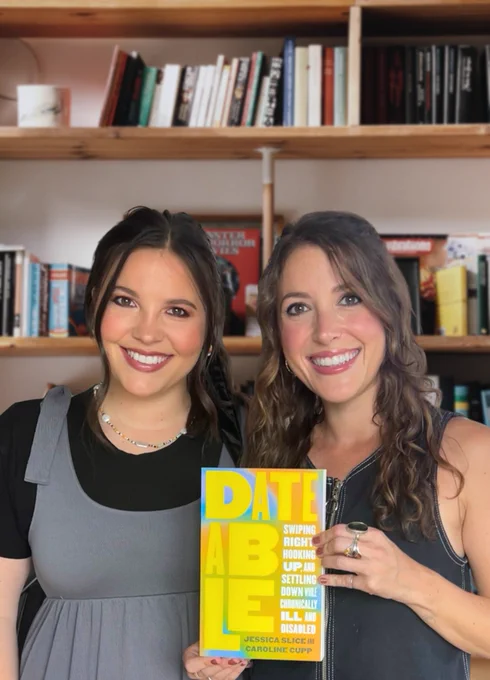 Dateability founders, Alexa and Jacqueline, two white brunette females in their 30s standing in front of a bookshelf. They are smiling and holding the book, Dateable, which has a colorful tie-dye background with the title in big block yellow letters.