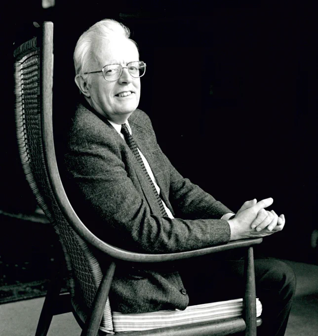 A black and white photo taken to the side of Sir David Cox in a wicker armchair, smiling at the camera with his hands clasped together in a relaxed manner in front of him while his his arms are on the arms of the chair.