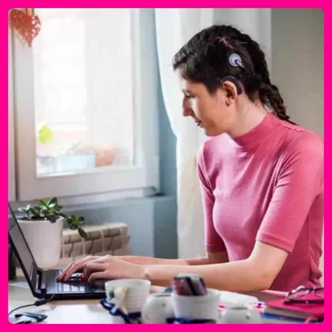 deaf female business executive using a cochlear implant processor on ear and magnetic skull headpiece that transmits external sounds to internal implant to assist in understanding speech and sound. She is wearing a pink turtleneck and working on her laptop in her office.   Photo Source:  Getty & Forbes 