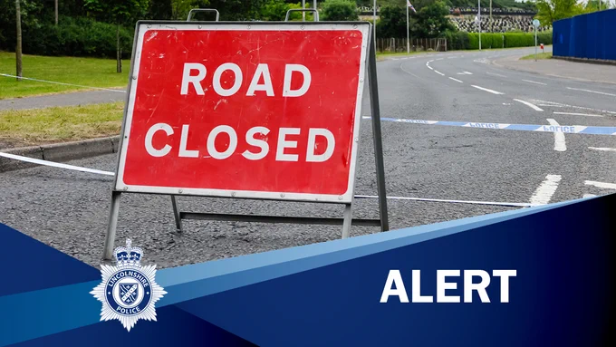 Red road closed sign placed on a road with blue and white cordon tape behind.  The word Alert appears in the bottom right hand corner. 