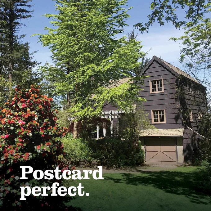 A rustic wooden house surrounded by lush trees and flowering bushes, with the text "Postcard perfect." overlaid on the image.