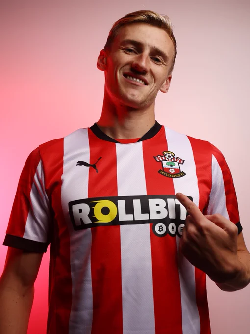 A photo of Flynn Downes pointing to the Southampton badge on his new PUMA red and white striped home kit, with a black collar and black ends to the sleeves, in front of a white background lit by a red light.