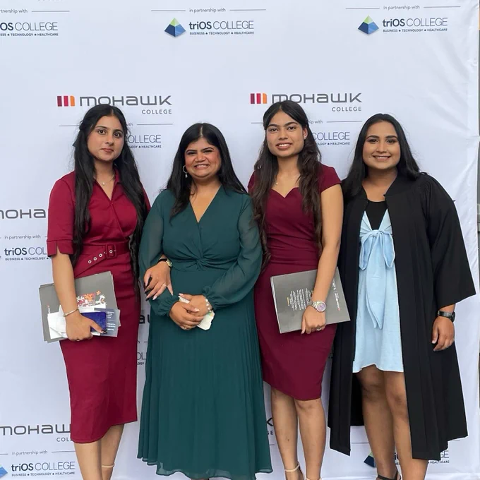 4 graduates standing in front of a Mohawk College and TriOS College backdrop, smiling at the camera.