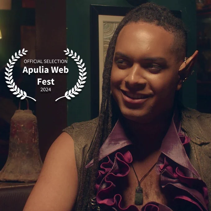 Uma foto de Inash na Taverna Original, sorrindo com malícia e olhando na direção dos louros de um festival, em que se lê: "Official Selection Apulia Web Fest 2024"