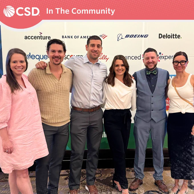 A photo of six people standing in a row smiling for the camera. A white backdrop is behind them with various logos. From Left to Right, a woman with a pink dress and long light brown hair, a man with medium wavy hair and goatee wearing a tan shirt and dress pants, a man with short brown hair wearing a ight blue dress buttoned shirt, dress pants and brown shoes, a woman with medium wavy brown hair with a white top and black pants and heels, a man with short hair wearing a dress shirt, bowtie, vest and a matching dress pants along with brown shoes, and lastly, a woman with brown hair in a bun, wearing glasses, white tank  top, black long skirt and dress flats. On the top is a red-orange border with white "CSD In The Community".
