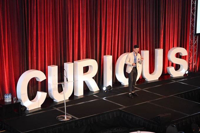 Nathan Kinch on stage in front of red curtains, and metre high white letters spelling Curious