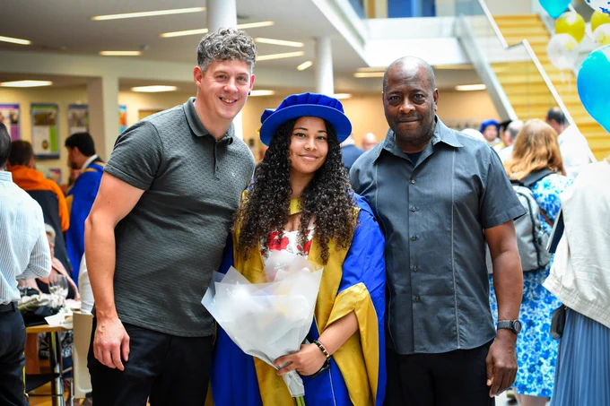 Person in postgraduate cap and gown celebrating graduation holding flowers with two men smiling either side