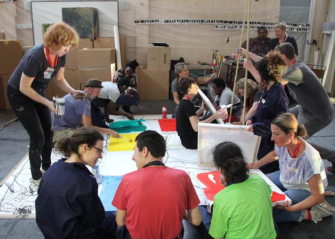 A group of people in a workshop. They are all painting canvases on the floor with bright colours and are happily conversing with each other.
