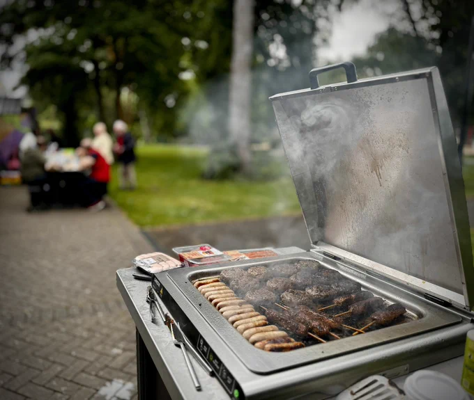 The lid on the public cooktop open, with burgers, sausages and kebabs cooking. Blurred in the background is a group of people gathered around a picnic table and eating.