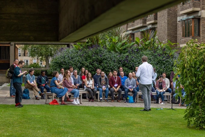 A group of people attentively listening to a speaker outdoors, with lush greenery in the background.