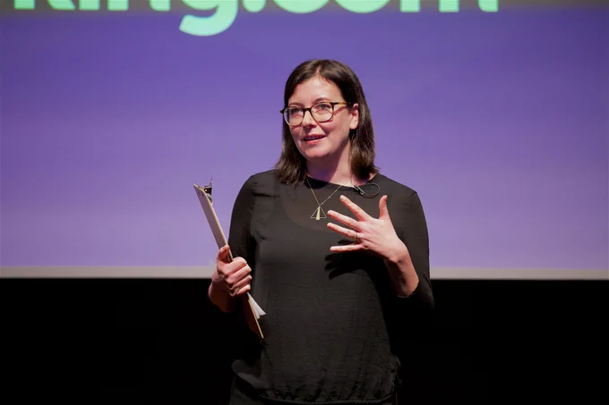 A person wearing glasses and a black outfit stands on a stage while holding a clipboard. They are gesturing with their other hand and are in front of a blue and purple screen.
