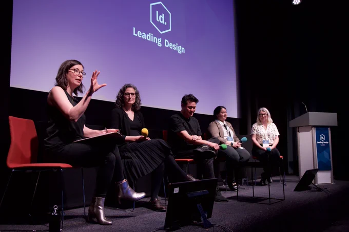 A group of five people seated on a stage, engaged in a panel discussion. The person on the far left is speaking animatedly with hand gestures. The backdrop displays the "Leading Design" logo. The setting appears to be a design conference.