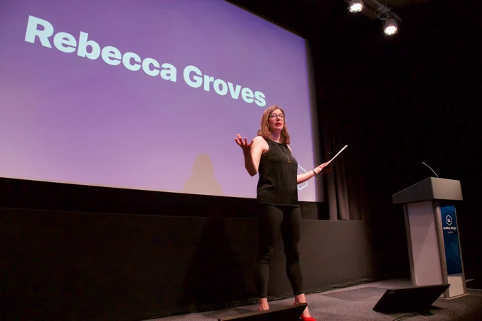 A person is standing on a stage with a microphone, speaking and gesturing with one hand while holding a piece of paper in the other. A presentation screen behind them displays the name "Rebecca Groves" on a purple background.