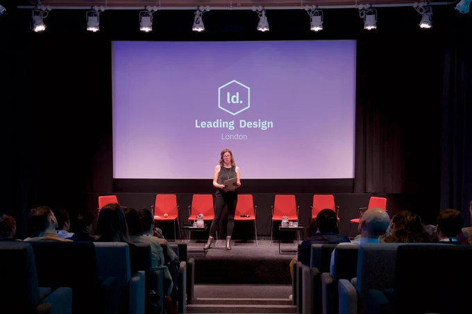 A person stands on a stage in front of a screen displaying "Leading Design London." The stage has five red chairs, and a seated audience is attentively watching. The venue appears to be a theater or conference space with a professional setting.