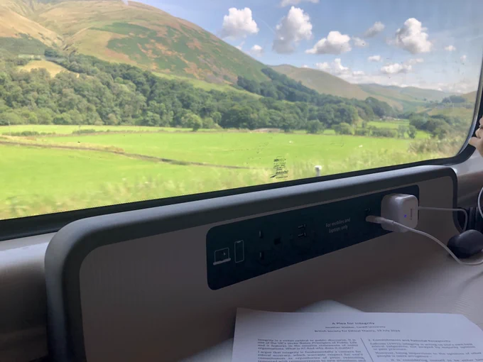 Photo of small mountains and blue skies through train window, with handout for my British Society for Ethical Theory talk on table in foreground.