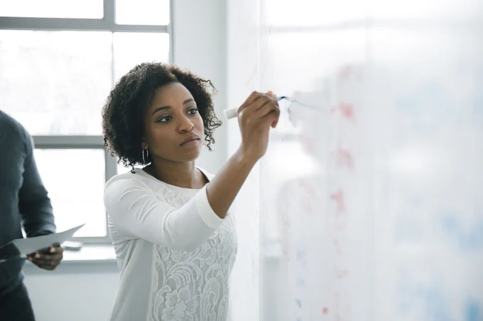 A person focused on writing on a whiteboard with an instructor standing behind them.