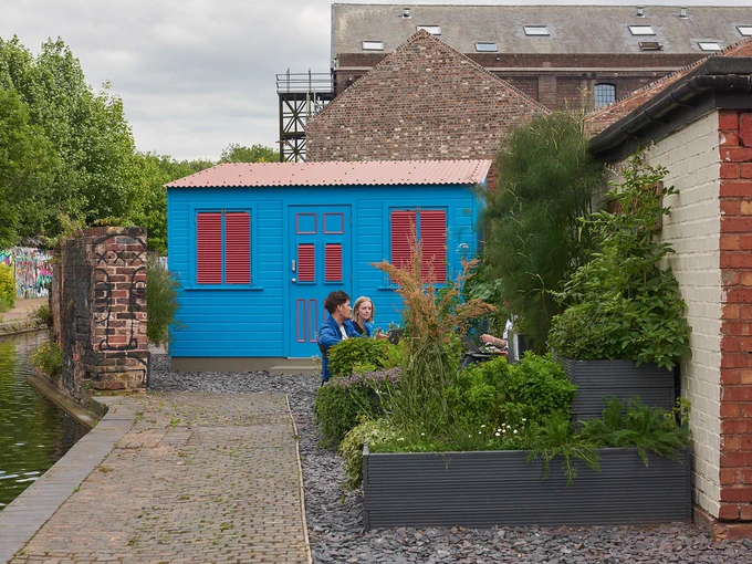 two people sit next to a bright blue shed in an urban garden next to a canal