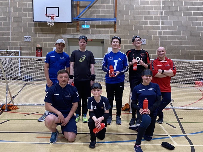 Cambridge Dons Aspire Day attendees including players and coaches with players holding their water bottles they received as a memento for attending the day. The photo is taken after the session in front of a goalball goal.
