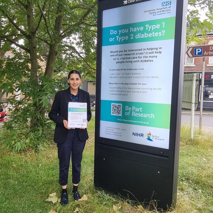Researcher Sam Hussain next to digital poster screen to #bepartofresearch on Normanton rd.