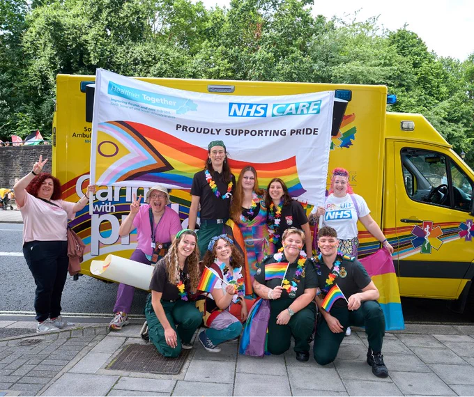 Bristol, North Somerset and South Gloucestershire healthcare staff holding a banner which says, 'NHS Care proudly supporting Pride' stood in front of a yellow ambulance. 