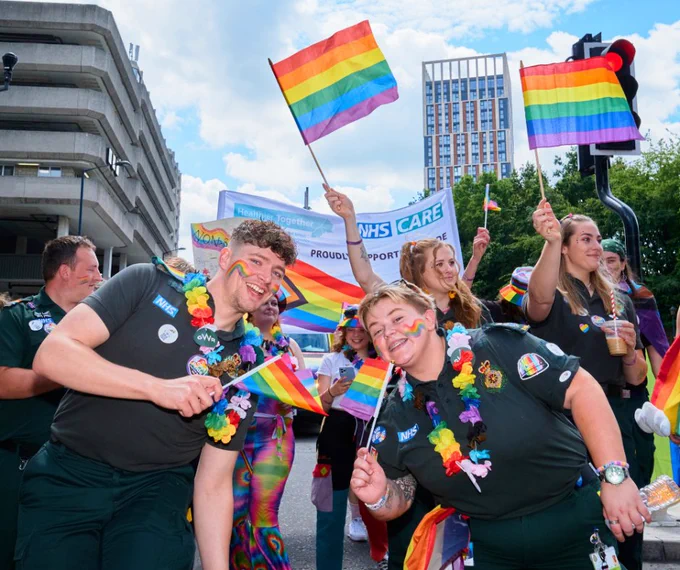 Bristol, North Somerset, and South Gloucestershire health care staff holding rainbow flags in front of a banner with NHS care written on it at Bristol Pride. 