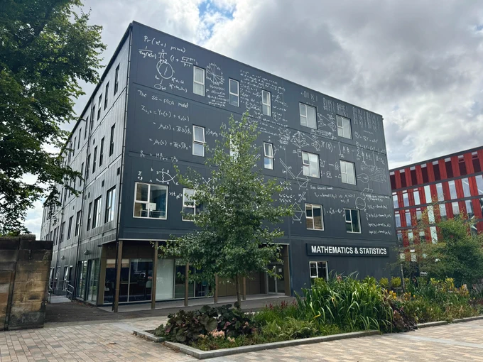 School of Mathematics & Statistics at the University of Glasgow covered in graphics. The white graphics contrast with the grey building and represent handwritten diagrams and equations from staff at the school that are used in their research. 

Trees and green plants are in front of the building, and to the right, the red School of Health and Wellbeing building can be seen in the background. There are cloudy skies with only slight blue skies visible between the clouds above the building. 