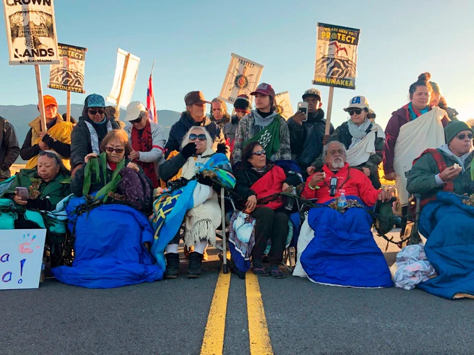 Photo by Caleb Jones with AP included in Business Insider article written by Joey Hadden entitled: "Photos show why many native Hawaiians don't want a giant telescope on the summit of Mauna Kea". The photo depicts numerous Indigenous elders (kūpuna) in the middle of Mauna Kea Access Road, seated with blankets in the early morning, with their supporters holding signs to block construction crews from ascending to the northern plateau of Mauna Kea to construct the Thirty Meter Telescope.
