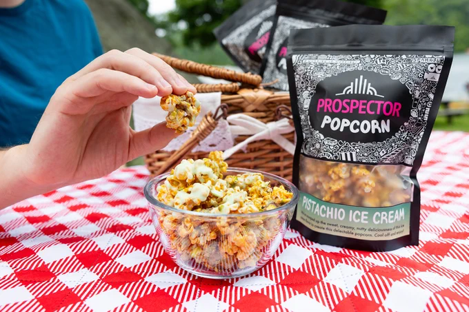 A bag of Pistachio Ice Cream sits on a red checked picnic blanket next to a bowl of its popcorn. A hand holds up a cluster of Pistachio Ice Cream gourmet popcorn. A picnic basket with more Prospector Popcorn bags can be seen in the background.