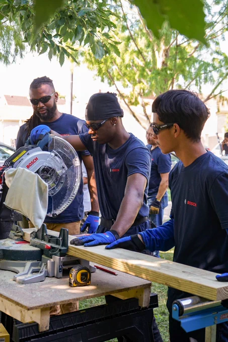 Three individuals using a miter saw to cut a wooden plank at an outdoor community service event. All are wearing protective gear and Bosch-branded shirts.