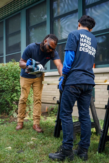 Two individuals engaged in outdoor work on a wooden structure near a building. One person is holding a power tool and focusing on making an adjustment. The other, wearing a shirt that reads "WHAT HARD WORKERS DESERVE," is observing. The setting includes a grassy area and a school building in the background.