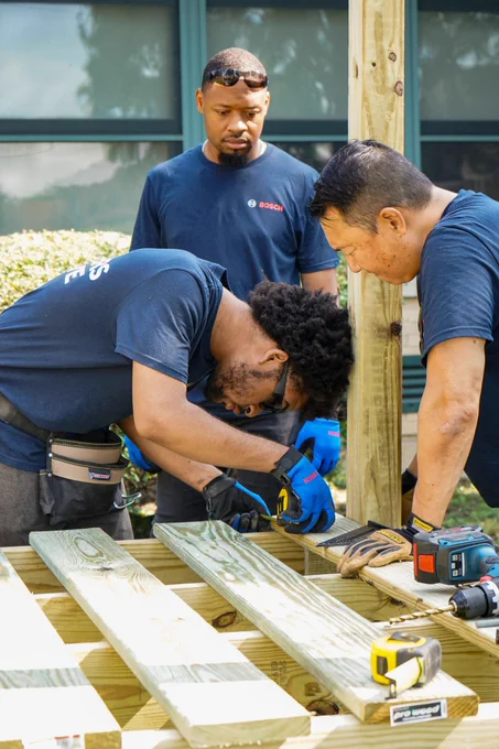 Three people working together on a carpentry project outdoors, with one measuring a wooden board as the others observe. They are surrounded by construction tools and materials.