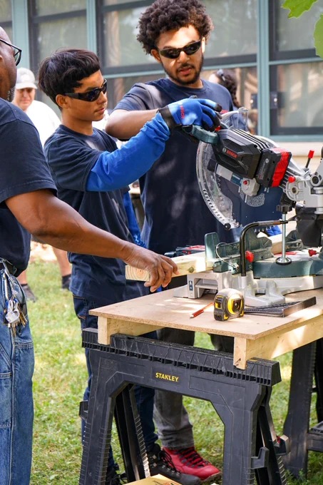 Two individuals using a compound miter saw on a workbench at an outdoor workshop, with another person observing. Tools and safety equipment visible.