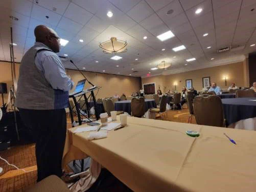 Leon T. Andrews, Jr. stands at a podium, speaking to a group of seated conference attendees.