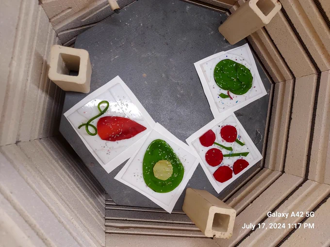 4 fruit-themed coasters in kiln, top view. Clickwise: green apple, 5 cherries, halved avocado, carrot, each with various stems and/or sparklies on top