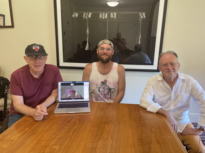 Matt sits in between Keith Tuma, who is wearing a red shirt, and Nik Money, who is wearing a white shirt. chris cheek attended virtually, and his face can be seen on a laptop screen, smiling and giving two thumbs up.