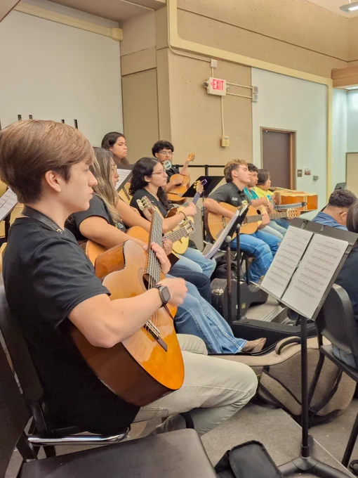 A group of students in a music classroom sits in rows, playing classical guitars and reading from music stands.