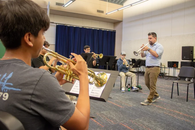 A music instructor is enthusiastically conducting a trumpet lesson with a group of students in a classroom setting, with chairs and music stands arranged around them.