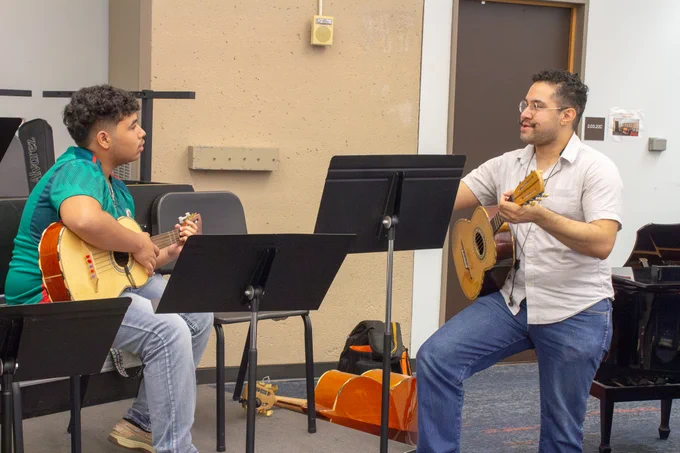 Two individuals seated with guitars in a music room, engaging in a guitar lesson. One is a young student holding an acoustic guitar, looking intently at the other, an instructor, who is demonstrating a chord on a smaller guitar. They are surrounded by music stands and other musical instruments.