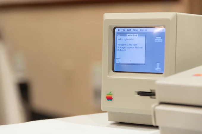 A tiny Macintosh 128K replica with an SD card inside the floppy drive. The small screen is showing the original Macintosh desktop with the Note Pad open. The Note Pad says, Hello, cyberpals! Welcome to the 18th Vintage Computer Festival Midwest.