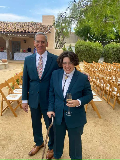 Dayn and their father, standing outside at a wedding venue wearing matching dark teal suits. Their father wears a floral tie and Dayn wears a bolo tie and holds a flute of champagne and a black cane