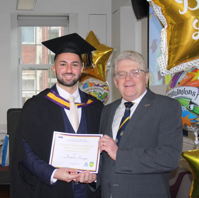 Arsallan and Liveryman Peter Reeve smiling together at the graduation celebrations. Arsallan is holding his prize certificate and wearing a graduation gown and mortarboard. There are balloons in the background which read: 'congratulations'.